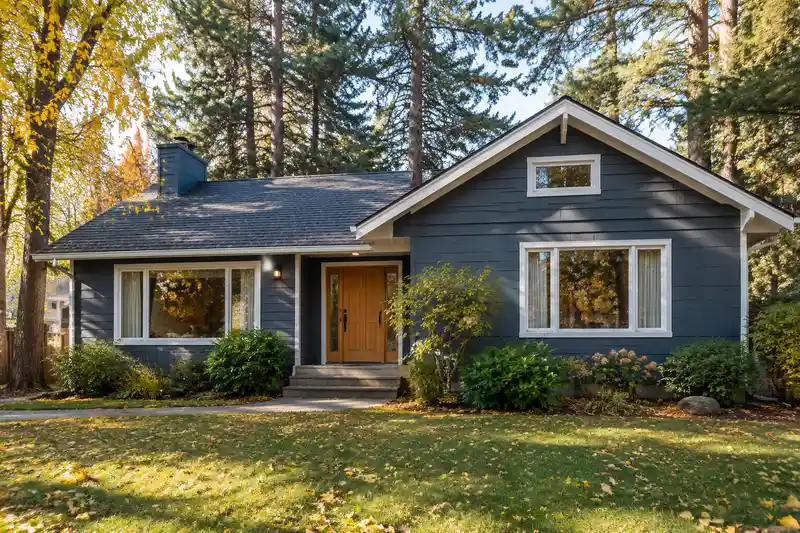 A cedar-sided home freshly painted deep blue-gray with white trim and a wood front door, under tall pines in early fall